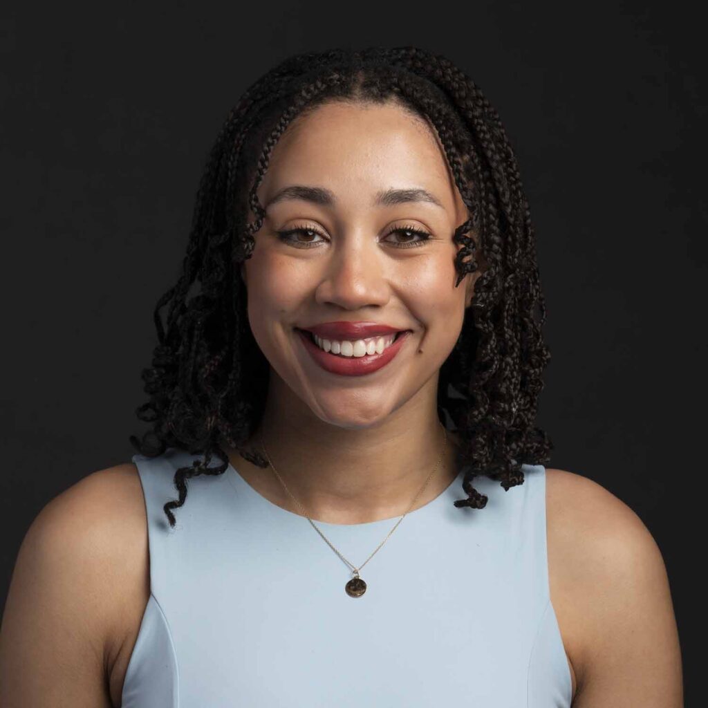 Smiling woman with braided hair wearing a light blue sleeveless top and gold necklace, posed against a dark background