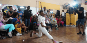 A dancer in a white outfit performing a dynamic floor dip on a wooden dance floor as a crowd cheers and a cameraman films.