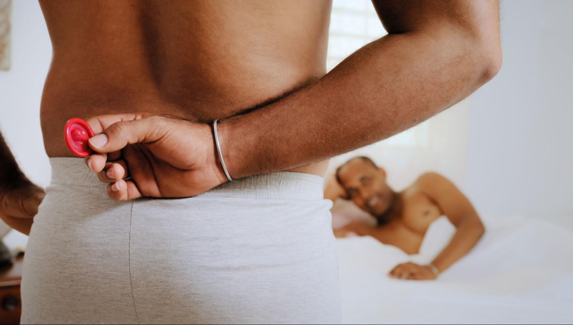Man holding condom behind back while male partner smiles in bed