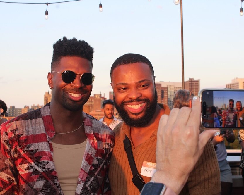 Two people smiling together on a rooftop at sunset while another person photographs them on a phone.