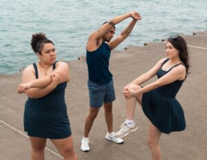 Three people in dark blue athletic wear stretching together on a concrete pier next to the water.