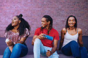 Three young people sitting outside against a brick wall, laughing while holding an ice cream cone and a blue speaker.